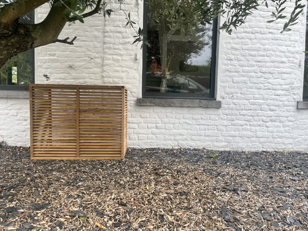 Cache de climatisation en teck Bokame contre une façade en briques peintes en blanc, sous un olivier